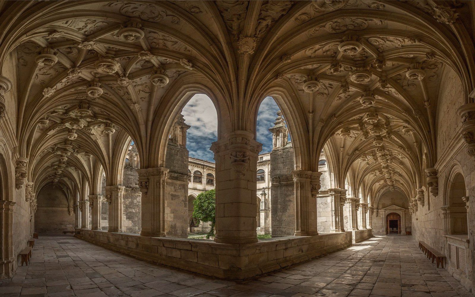 Ornate ribbed vaulting arches over a stone cloister walkway surrounding an open courtyard garden